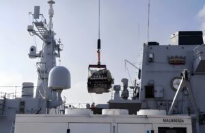 Crane on a gray warship hoists a heavy engine or equipment over the deck.