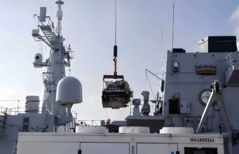 Crane on a gray warship hoists a heavy engine or equipment over the deck.