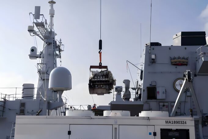 Crane on a gray warship hoists a heavy engine or equipment over the deck.