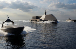 A large, angular gray naval vessel sails the open sea, with two smaller speedboats nearby and white wake in the water.