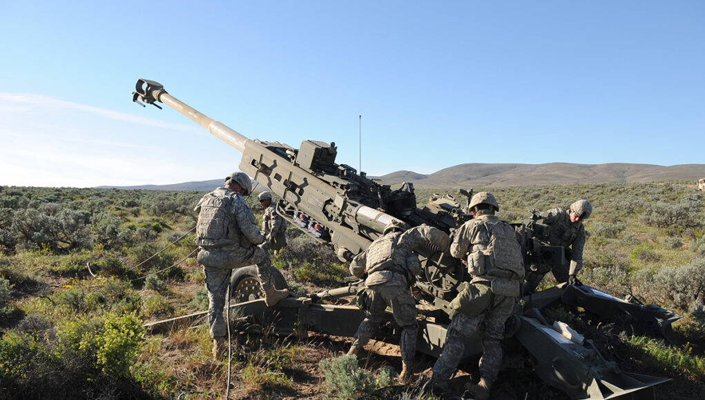 Soldiers in camouflage work around a large field artillery gun in a desert training area.