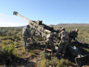Soldiers in camouflage work around a large field artillery gun in a desert training area.