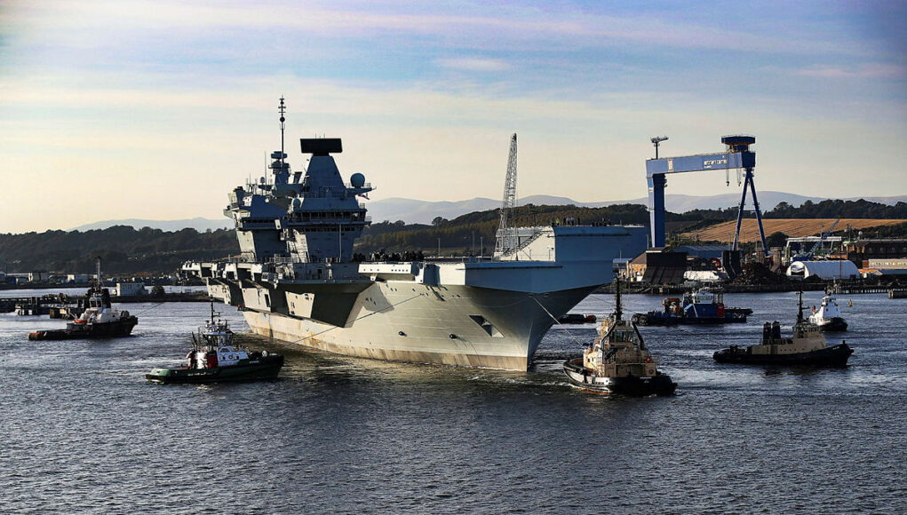 Large gray aircraft carrier moored in a harbor, surrounded by tugboats and industrial cranes in the background.