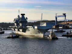 Large gray aircraft carrier moored in a harbor, surrounded by tugboats and industrial cranes in the background.