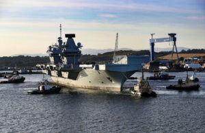 Large gray aircraft carrier moored in a harbor, surrounded by tugboats and industrial cranes in the background.