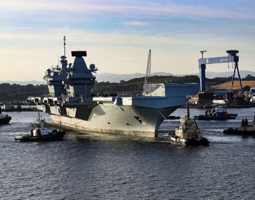 Large gray aircraft carrier moored in a harbor, surrounded by tugboats and industrial cranes in the background.