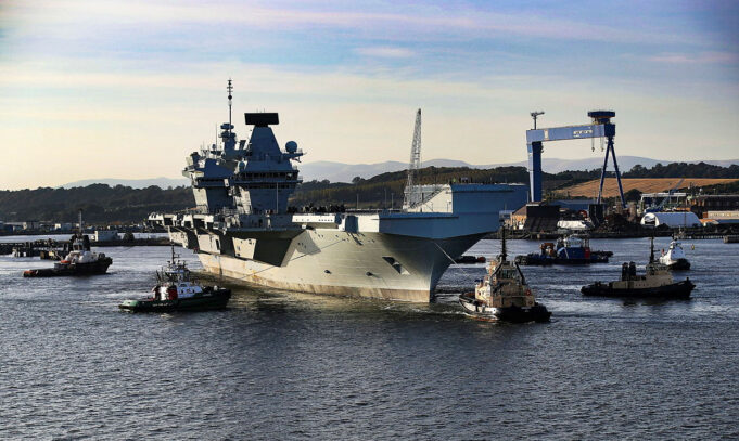 Large gray aircraft carrier moored in a harbor, surrounded by tugboats and industrial cranes in the background.