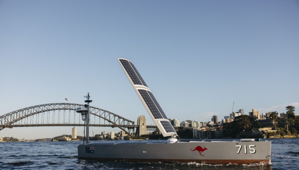 Ocius Bluebottle in Sydney Harbour 3 Solar-powered boat with a large solar sail, number 715 and a red kangaroo logo, cruising near Sydney Harbour Bridge with a city skyline in the background.