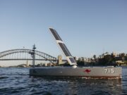 Uncrewed Bluebottle vessels push persistent maritime surveillance Solar-powered boat with a large solar sail, number 715 and a red kangaroo logo, cruising near Sydney Harbour Bridge with a city skyline in the background.