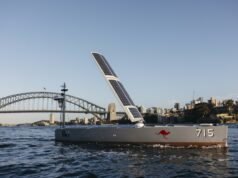 Solar-powered boat with a large solar sail, number 715 and a red kangaroo logo, cruising near Sydney Harbour Bridge with a city skyline in the background.