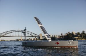 Solar-powered boat with a large solar sail, number 715 and a red kangaroo logo, cruising near Sydney Harbour Bridge with a city skyline in the background.