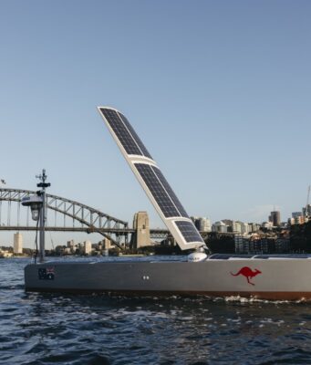 Bluebottle maker opens Sydney plant for forty vessels Solar-powered boat with a large solar sail, number 715 and a red kangaroo logo, cruising near Sydney Harbour Bridge with a city skyline in the background.