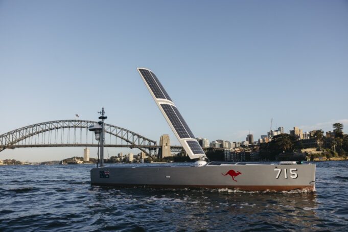 Solar-powered boat with a large solar sail, number 715 and a red kangaroo logo, cruising near Sydney Harbour Bridge with a city skyline in the background.