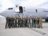 Group of military crew in green flight suits standing in front of a large white Marine aircraft on a tarmac with stairs deployed, posing for a photo.