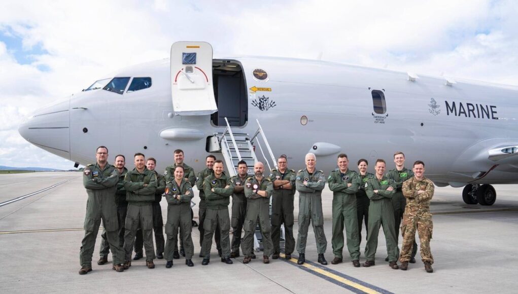 Group of military crew in green flight suits standing in front of a large white Marine aircraft on a tarmac with stairs deployed, posing for a photo.