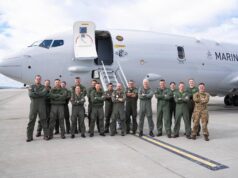 Group of military crew in green flight suits standing in front of a large white Marine aircraft on a tarmac with stairs deployed, posing for a photo.