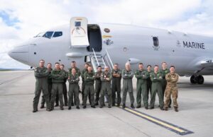 Group of military crew in green flight suits standing in front of a large white Marine aircraft on a tarmac with stairs deployed, posing for a photo.