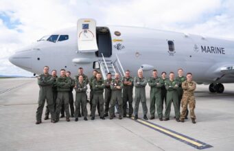 Group of military crew in green flight suits standing in front of a large white Marine aircraft on a tarmac with stairs deployed, posing for a photo.