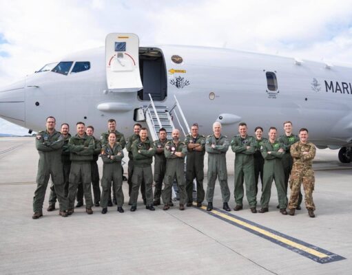 Group of military crew in green flight suits standing in front of a large white Marine aircraft on a tarmac with stairs deployed, posing for a photo.