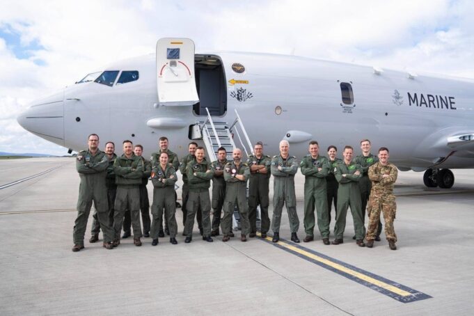 Group of military crew in green flight suits standing in front of a large white Marine aircraft on a tarmac with stairs deployed, posing for a photo.