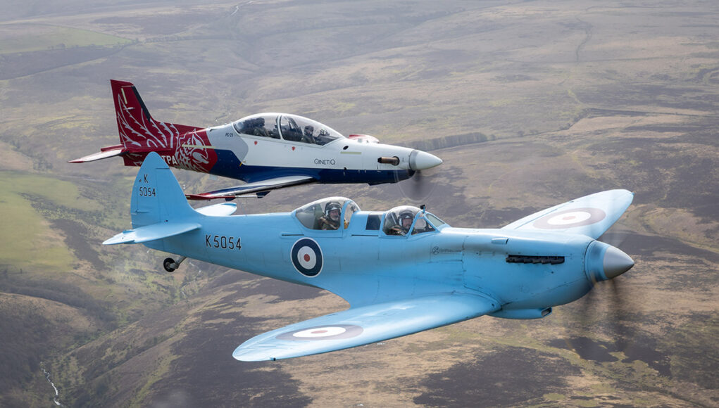 Two vintage RAF training aircraft fly in formation over rolling countryside, one blue with roundels in the foreground and a red‑white aircraft behind it.