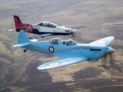 Two vintage RAF training aircraft fly in formation over rolling countryside, one blue with roundels in the foreground and a red‑white aircraft behind it.