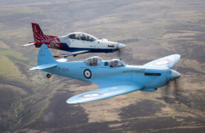 Two vintage RAF training aircraft fly in formation over rolling countryside, one blue with roundels in the foreground and a red‑white aircraft behind it.