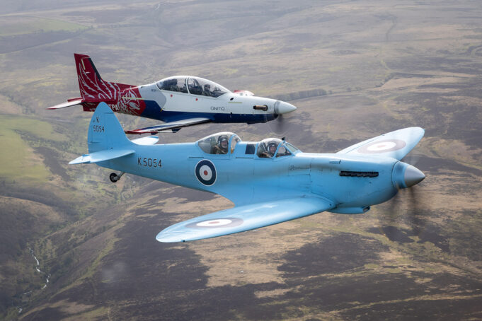 Two vintage RAF training aircraft fly in formation over rolling countryside, one blue with roundels in the foreground and a red‑white aircraft behind it.