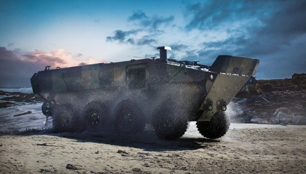 Armored amphibious vehicle driving on a wet sandy beach as seawater sprays from its wheels at the shore.