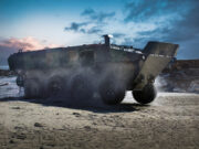 Armored amphibious vehicle driving on a wet sandy beach as seawater sprays from its wheels at the shore.
