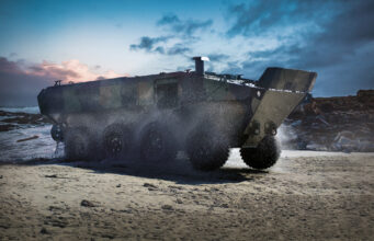 Armored amphibious vehicle driving on a wet sandy beach as seawater sprays from its wheels at the shore.