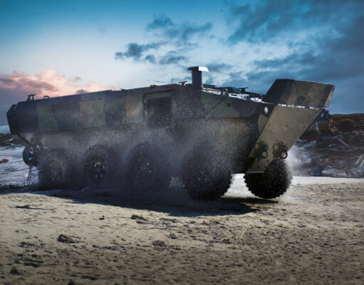 Armored amphibious vehicle driving on a wet sandy beach as seawater sprays from its wheels at the shore.