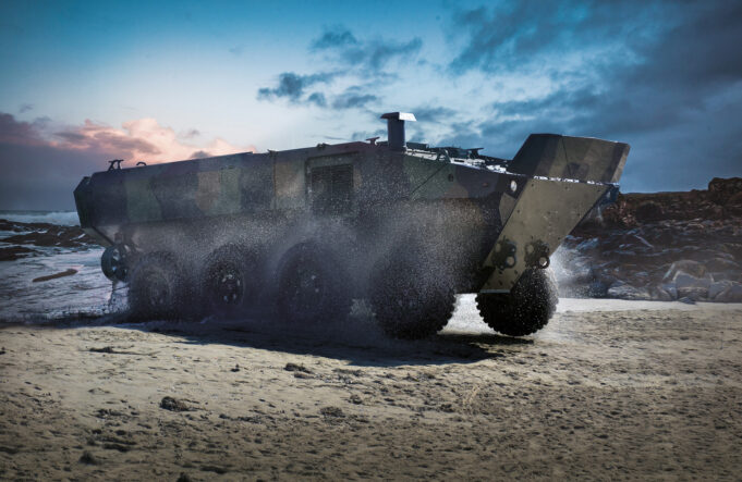 Armored amphibious vehicle driving on a wet sandy beach as seawater sprays from its wheels at the shore.