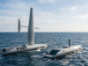 Two autonomous saildrone vessels ride the open ocean, one with a tall mast and a horizontal sail and the other nearby, horizon in the distance.