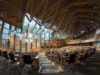Large modern parliamentary chamber with curved wood benches and black office chairs in rows.