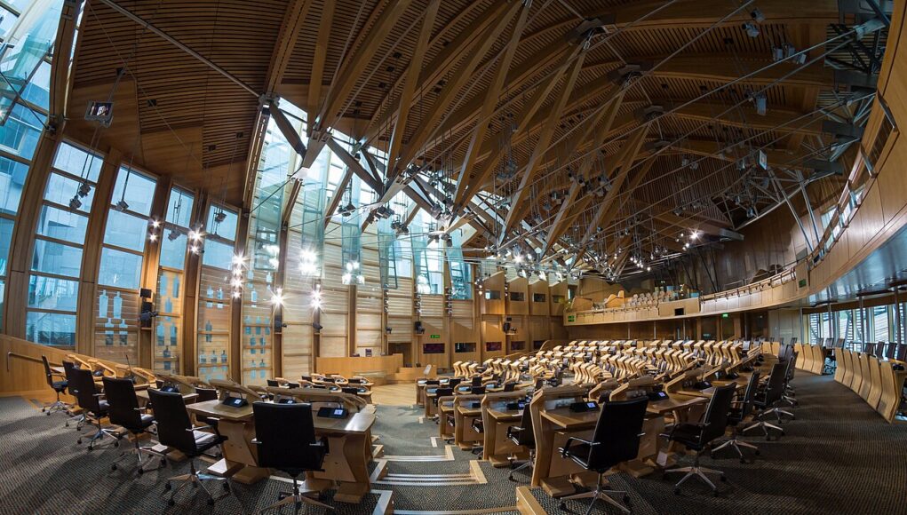 Large modern parliamentary chamber with curved wood benches and black office chairs in rows.
