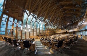 Large modern parliamentary chamber with curved wood benches and black office chairs in rows.