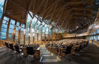 Large modern parliamentary chamber with curved wood benches and black office chairs in rows.