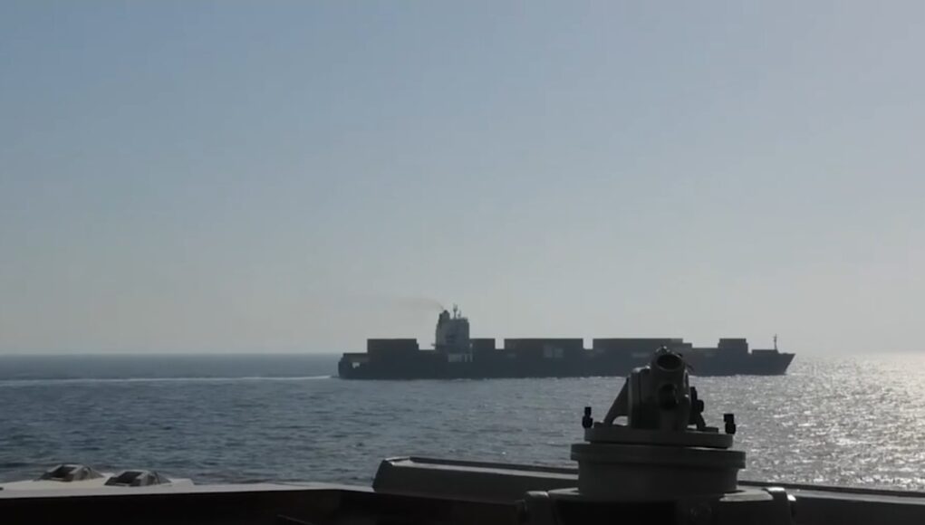View from a warship deck: a forward gun mount in the foreground with a distant large carrier-like vessel on the calm sea at sunset/light glare on the water.