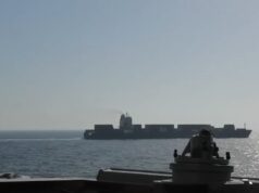 View from a warship deck: a forward gun mount in the foreground with a distant large carrier-like vessel on the calm sea at sunset/light glare on the water.
