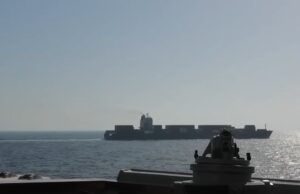 View from a warship deck: a forward gun mount in the foreground with a distant large carrier-like vessel on the calm sea at sunset/light glare on the water.