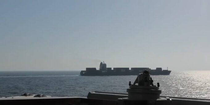 View from a warship deck: a forward gun mount in the foreground with a distant large carrier-like vessel on the calm sea at sunset/light glare on the water.