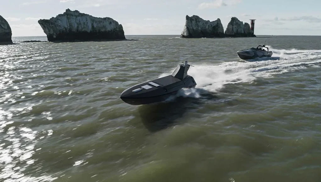 Two black speedboats race across choppy green water near rugged rocky islets with a distant red-and-white lighthouse on the horizon.