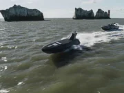 Two black speedboats race across choppy green water near rugged rocky islets with a distant red-and-white lighthouse on the horizon.