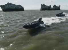 Two black speedboats race across choppy green water near rugged rocky islets with a distant red-and-white lighthouse on the horizon.