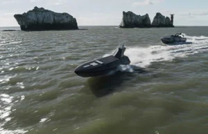 Two black speedboats race across choppy green water near rugged rocky islets with a distant red-and-white lighthouse on the horizon.