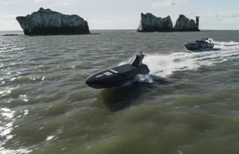 Two black speedboats race across choppy green water near rugged rocky islets with a distant red-and-white lighthouse on the horizon.