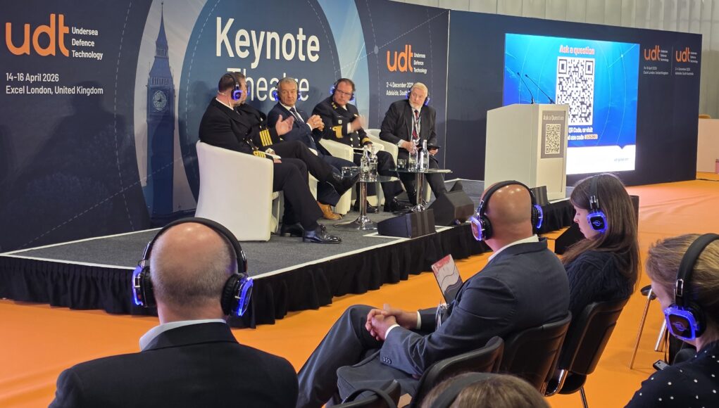 Panel discussion on stage with five men in suits and uniforms seated in white chairs at a UDT Keynote Theatre event, audience wearing blue headphones on orange floor.