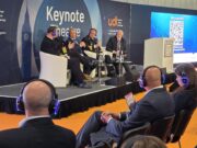 Panel discussion on stage with five men in suits and uniforms seated in white chairs at a UDT Keynote Theatre event, audience wearing blue headphones on orange floor.
