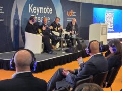 Panel discussion on stage with five men in suits and uniforms seated in white chairs at a UDT Keynote Theatre event, audience wearing blue headphones on orange floor.